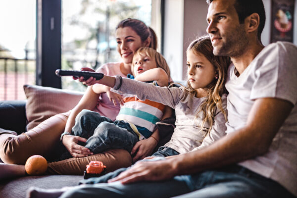 Young happy family watching TV on sofa at home. Focus is on girl changing channels.