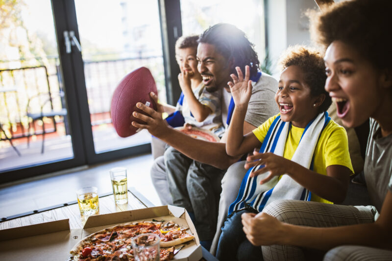 Young joyful black family having fun while cheering for their favorite American football team at home.