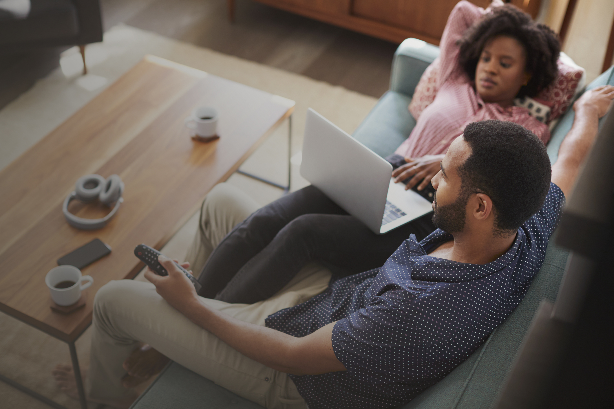 Couple Sitting On Sofa At Home Using Laptop Computer And Watching TV